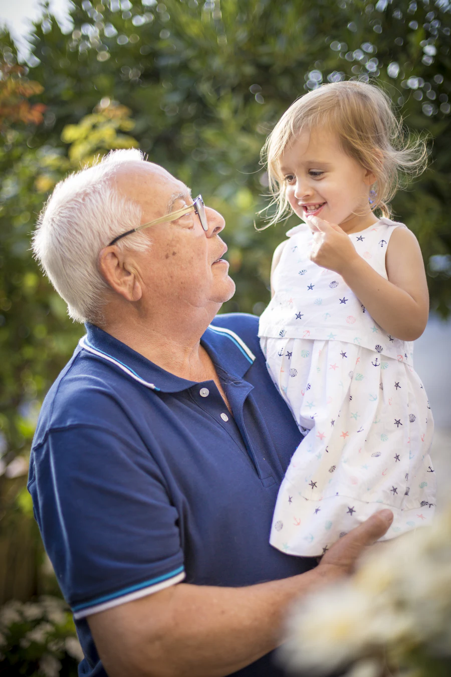 A grandfather holding his young granddaughter; both smiling at each other in a garden.