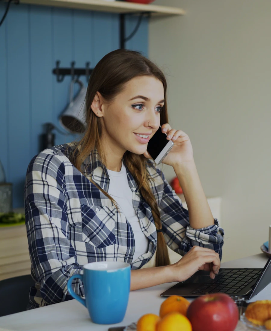 A woman at her kitchen table on a phone call, typing on her laptop.
