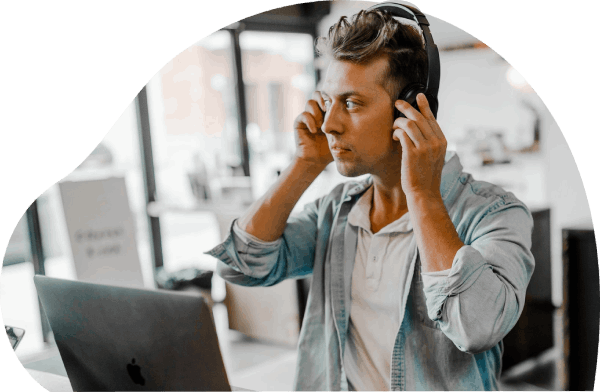 A curvy-cropped image of a man in his late 20s with blonde hair, gently adjusting his wireless headphones with both his hands, while looking away from his Apple computer.