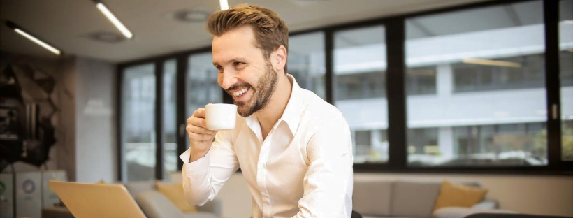 A person sitting in a co-working-like space, drinking his coffee and smiling at someone or something.